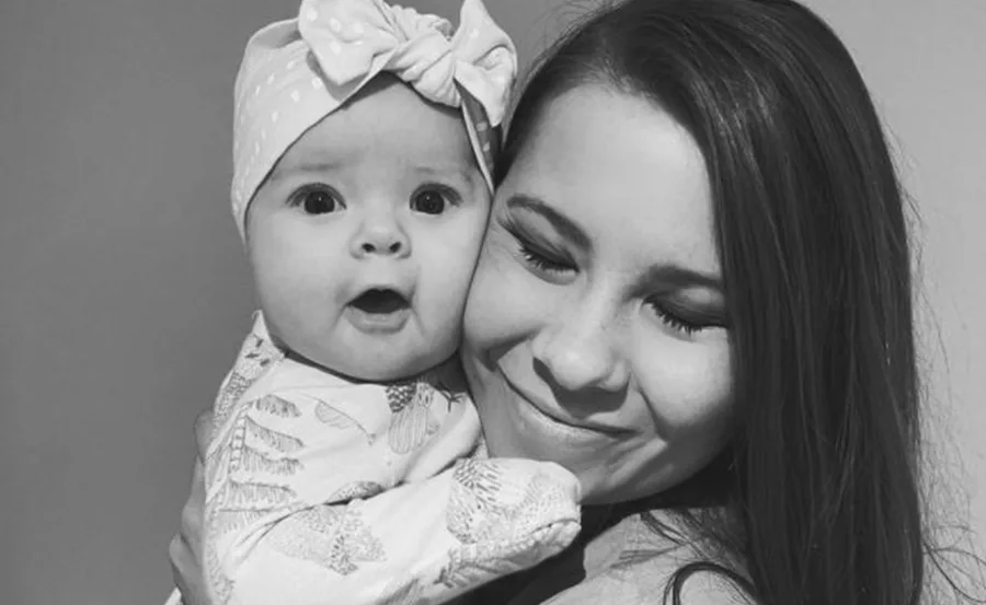 A woman smiling with eyes closed, holding a baby with a headband, both in black-and-white close-up.