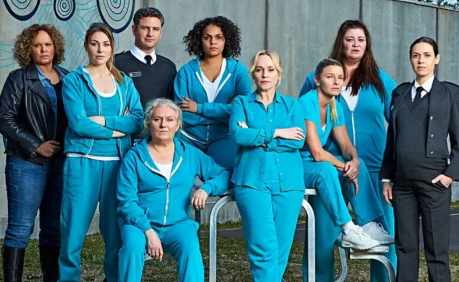 Cast of "Wentworth" in blue uniforms and guards posing together against a concrete wall backdrop.