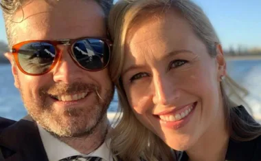Two people smiling outdoors, one wearing sunglasses, with water in the background.