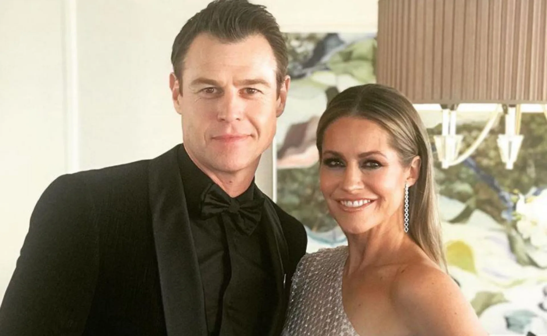 Man in a tuxedo and woman in a sequined dress smiling, standing in a well-lit room.