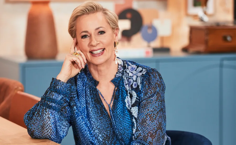 A person with short blonde hair smiles, wearing a blue floral-patterned blouse, seated indoors.