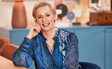 A person with short blonde hair smiles, wearing a blue floral-patterned blouse, seated indoors.
