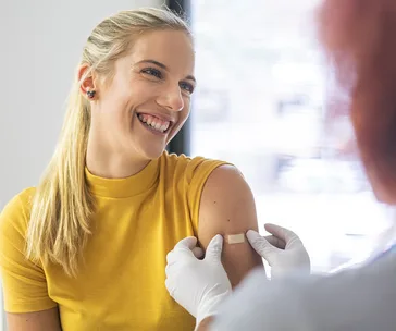 Smiling woman in yellow shirt receives a flu shot with a bandage on her arm in a healthcare setting.