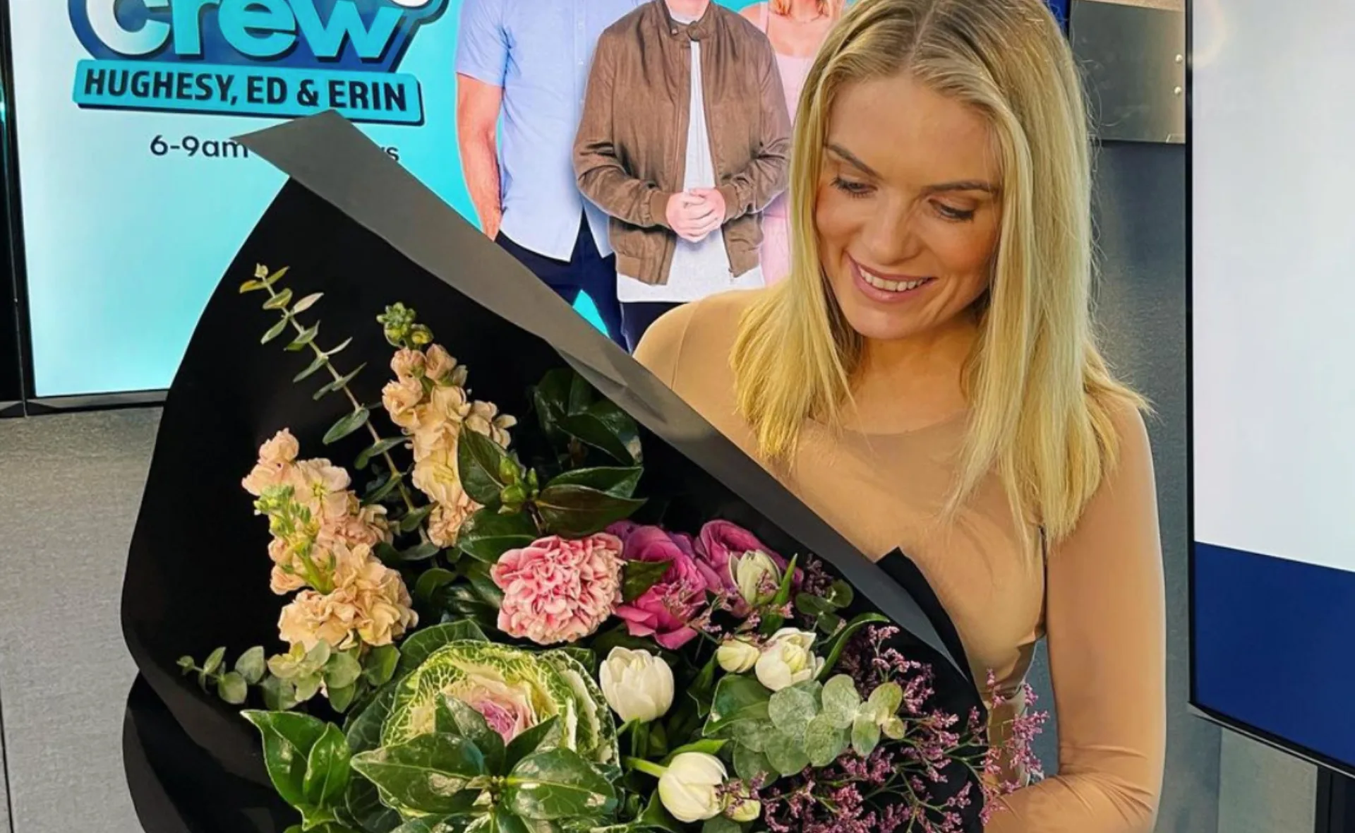 Woman smiling at a bouquet of flowers, with a radio show poster in the background.