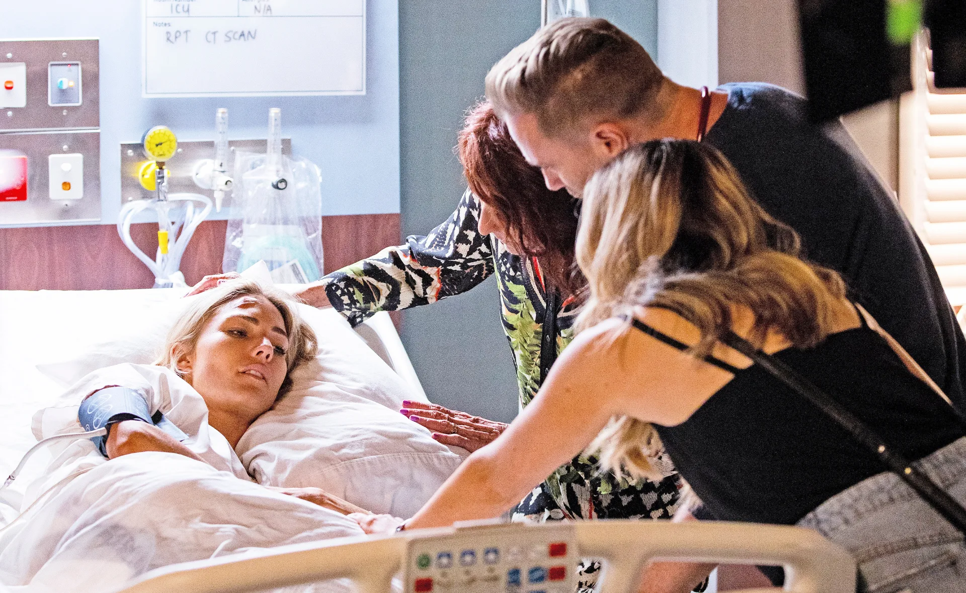 A woman lying in a hospital bed surrounded by three people, one holding her hand, looking concerned.