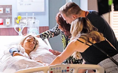 A woman lying in a hospital bed surrounded by three people, one holding her hand, looking concerned.