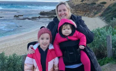 A woman with two young children, one in a pink jacket, standing on a beach with ocean in the background.