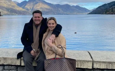 A couple sits by a lake with mountains in the background, smiling at the camera.
