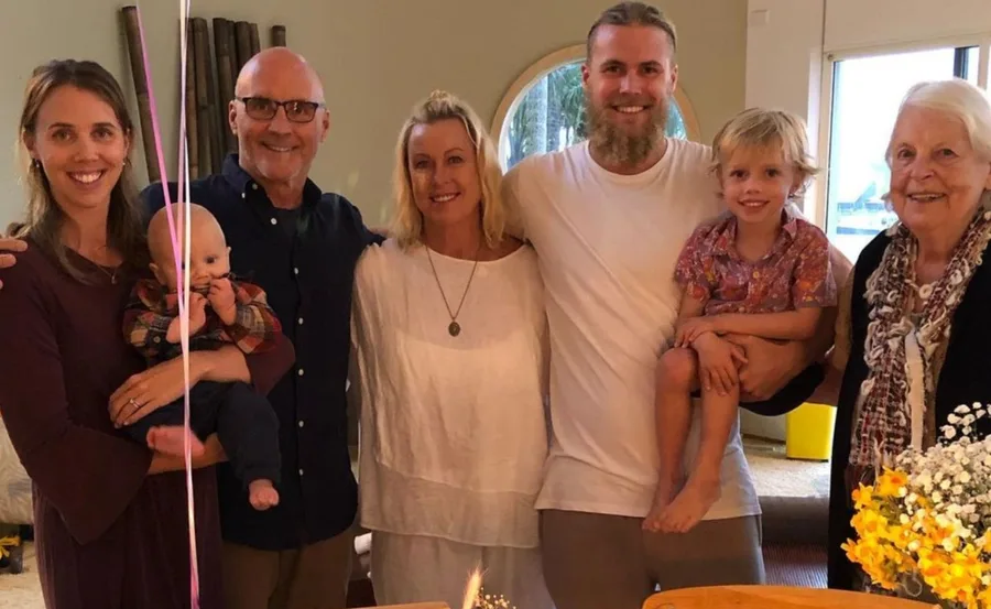 A smiling family gathered together indoors, with a baby and a child, celebrating a special occasion near a table with flowers.