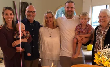 A smiling family gathered together indoors, with a baby and a child, celebrating a special occasion near a table with flowers.