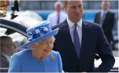 Queen in a blue outfit with hat and brooch smiles beside a man in a suit outdoors, near a car.