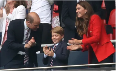 A young boy in a suit claps enthusiastically, flanked by two adults applauding at a football match.