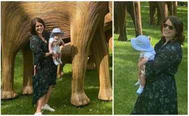 Woman holding a baby, standing beside large wooden elephant sculptures in a grassy park.