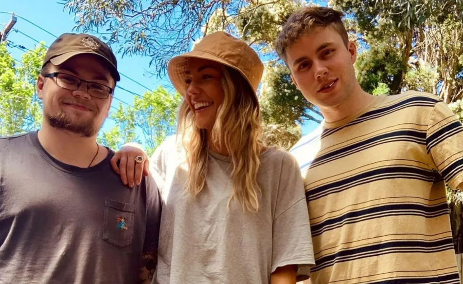 Three people smiling outdoors, with trees in the background; one wears a hat, another a striped shirt.