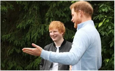Two individuals standing outdoors, one gesturing while the other smiles, with greenery in the background.