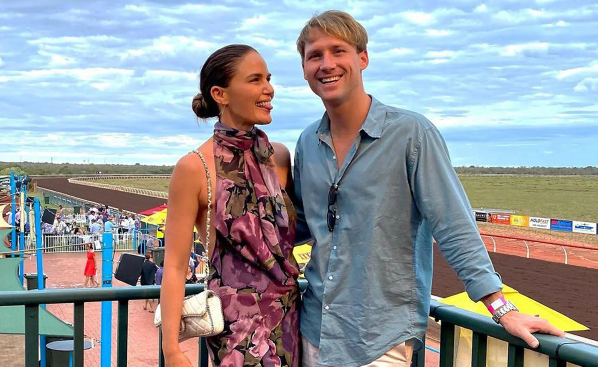 A couple stands smiling at a racecourse, the woman in a floral dress and the man in a blue shirt.
