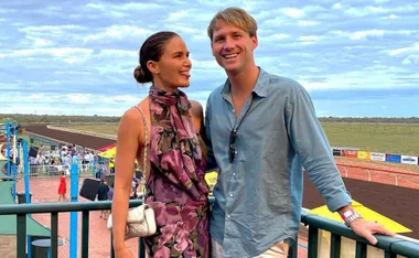 A couple stands smiling at a racecourse, the woman in a floral dress and the man in a blue shirt.