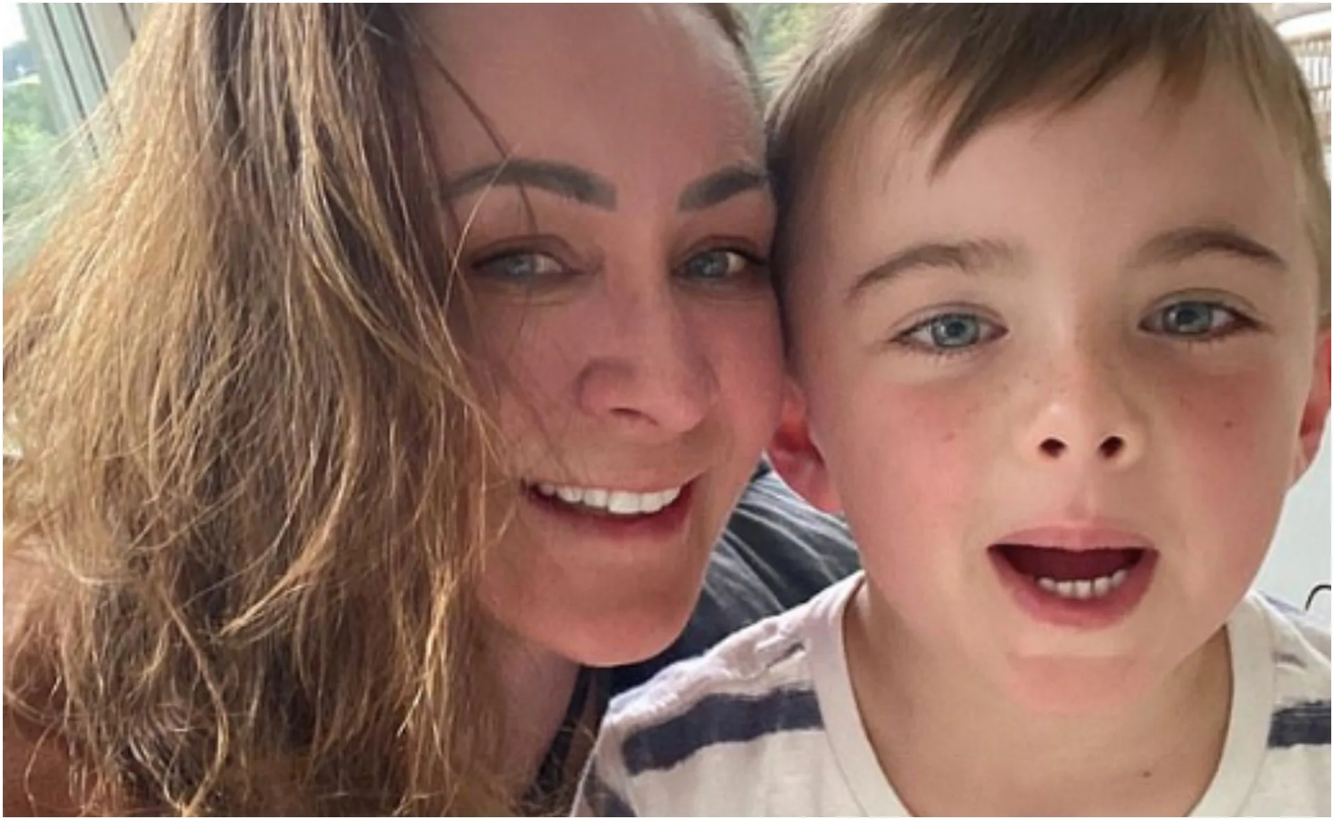 A woman with long hair smiles beside a young boy with short hair and a striped shirt, both looking at the camera.