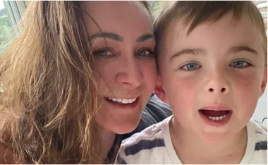 A woman with long hair smiles beside a young boy with short hair and a striped shirt, both looking at the camera.