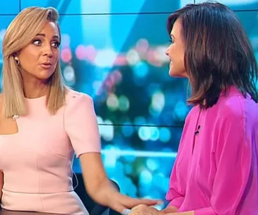 Two women in conversation on a TV set with blurred city lights background.