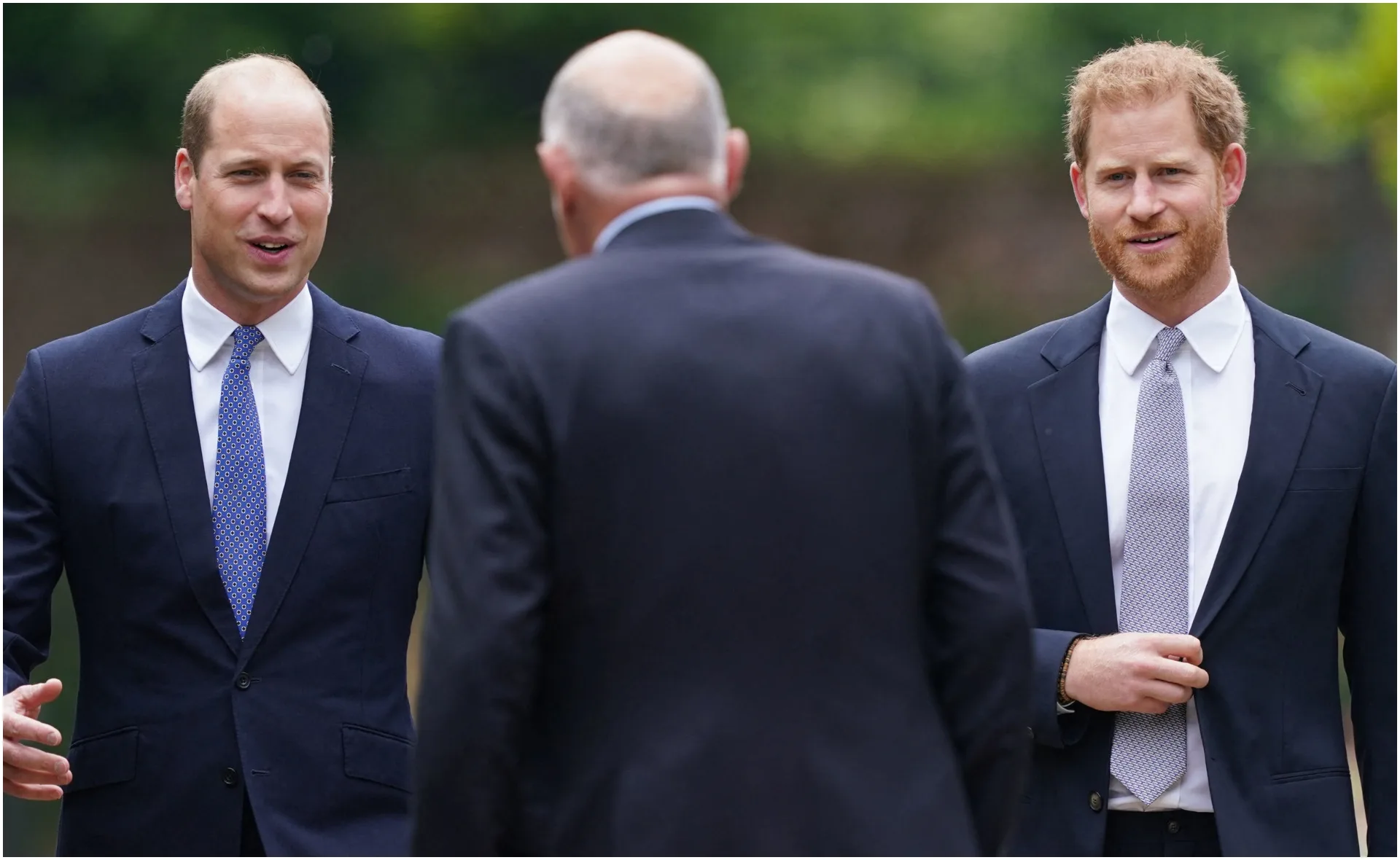 Two men in suits smiling outdoors, engaging in conversation with a bald man facing away.