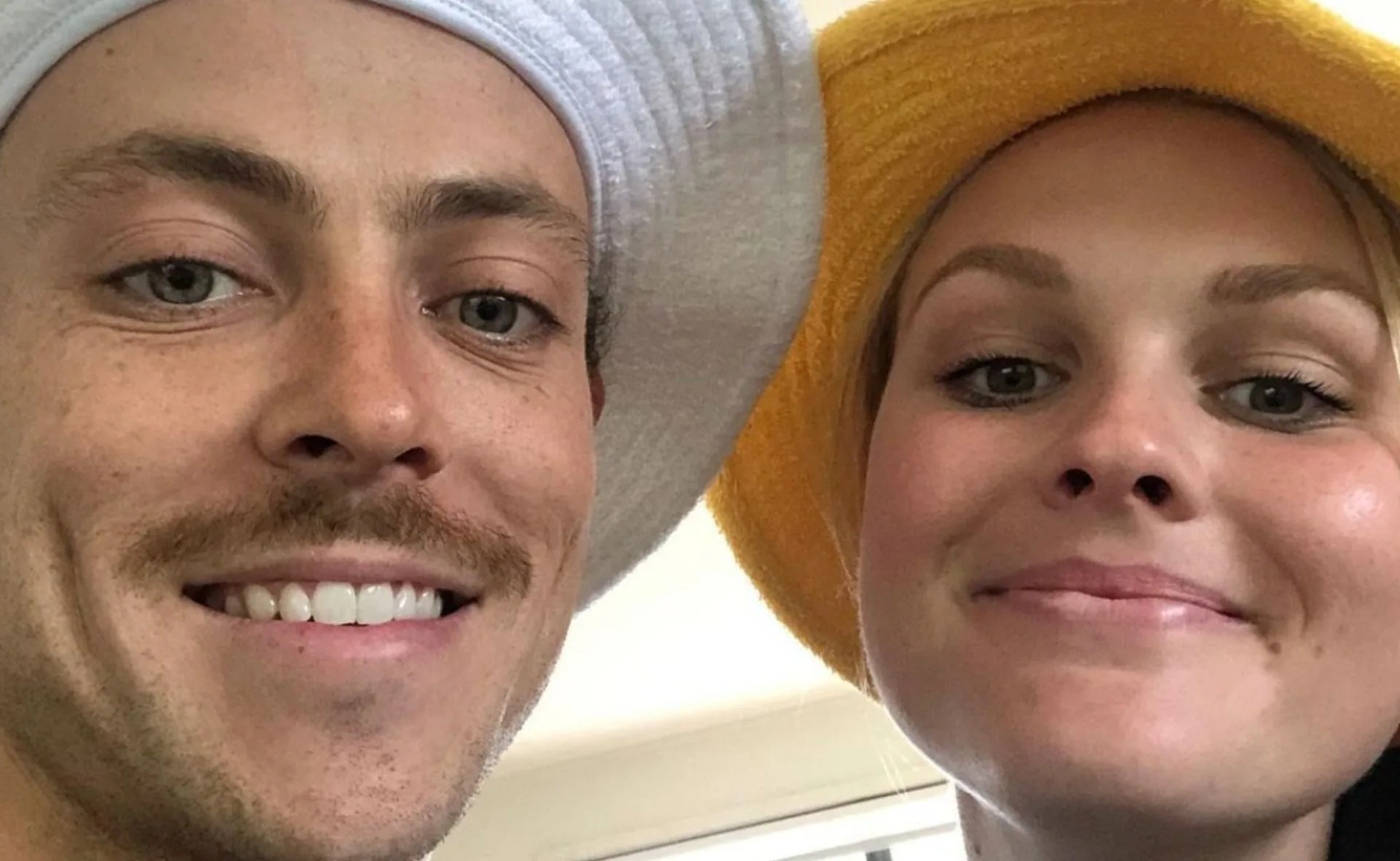 Two people smiling, wearing bucket hats—one white, one yellow—posing closely for a selfie indoors.