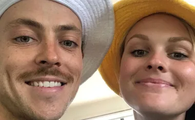 Two people smiling, wearing bucket hats—one white, one yellow—posing closely for a selfie indoors.