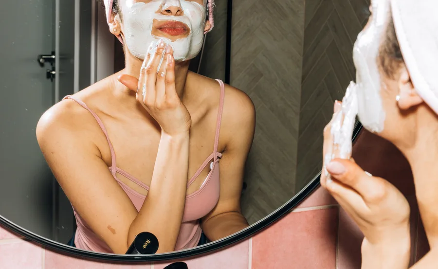 Woman applying a white facial mask in front of a mirror, wearing a pink tank top and headband.