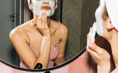Woman applying a white facial mask in front of a mirror, wearing a pink tank top and headband.