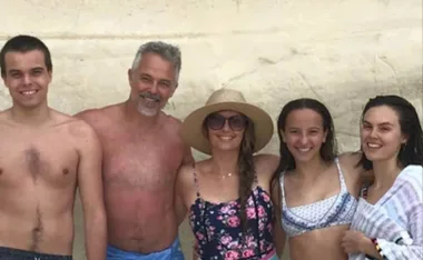 A smiling group of five people at the beach, with one wearing a sun hat and floral dress.