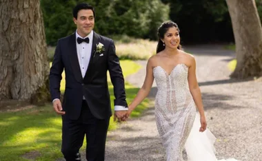 A couple walking hand in hand on a garden path; the woman in a strapless wedding dress, the man in a tuxedo.