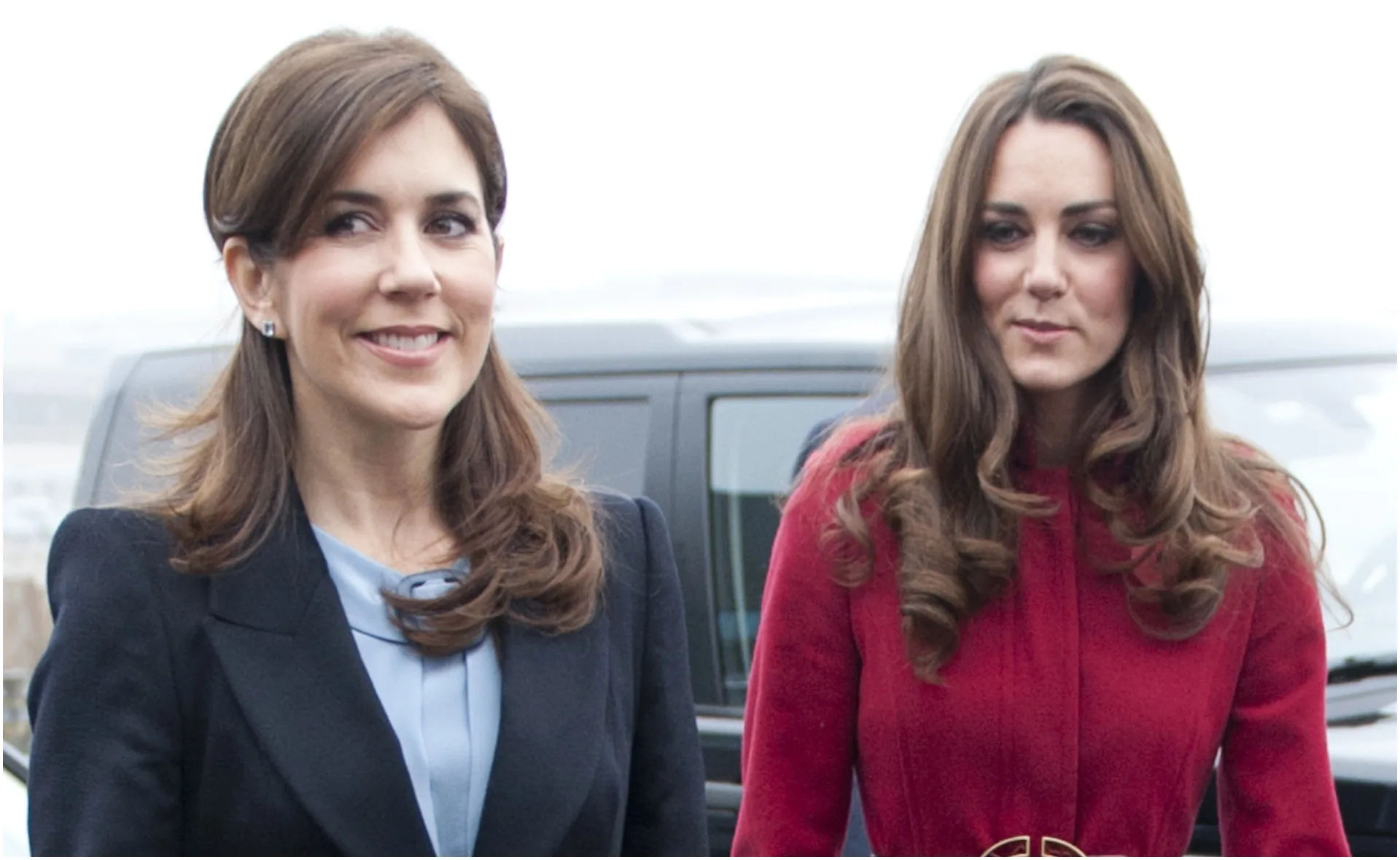 Two women with long brown hair, one in a black coat and one in a red coat, standing outdoors near a vehicle.