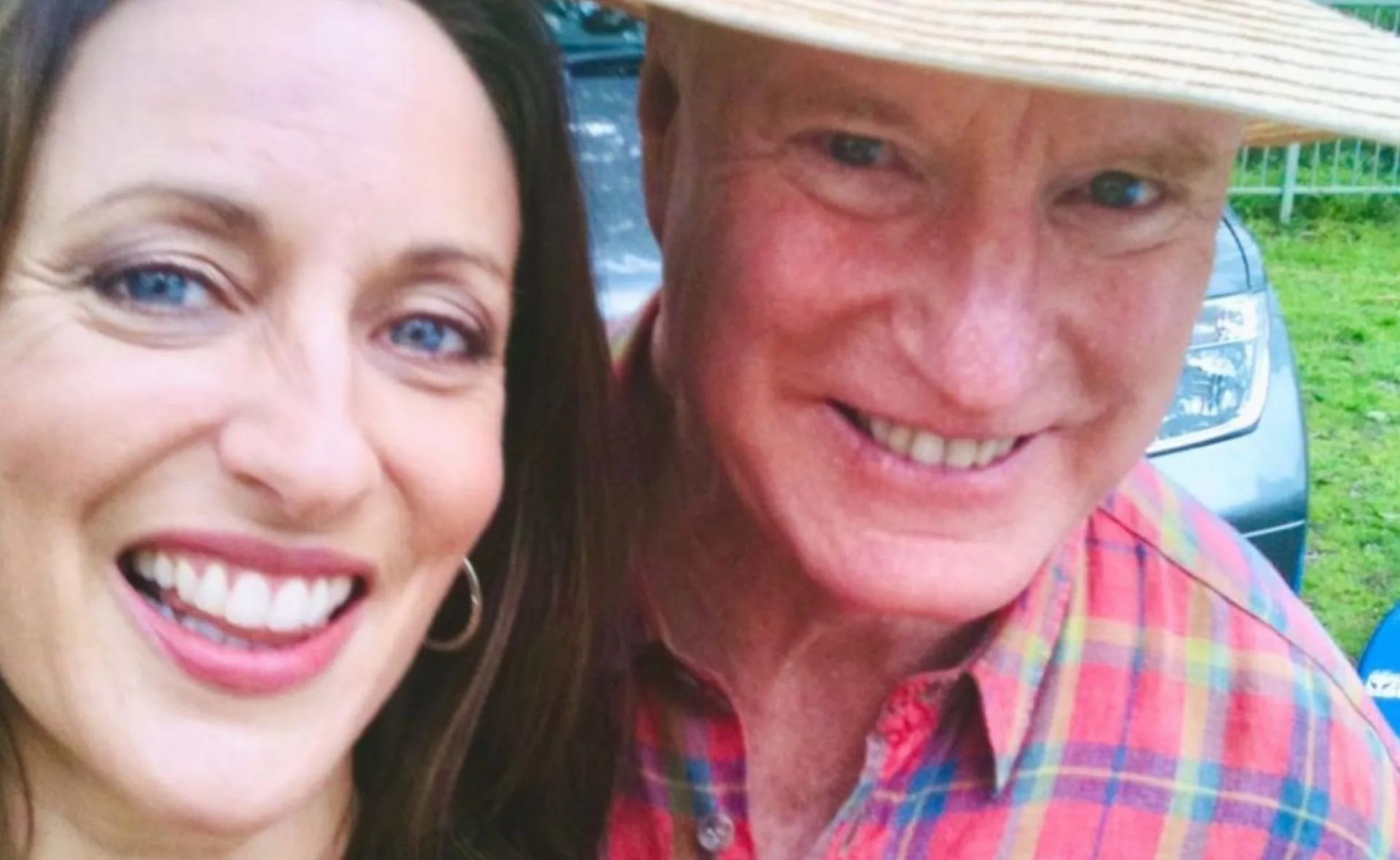 Smiling duo take a selfie outdoors, one wears a straw hat and plaid shirt, the other has hoop earrings.