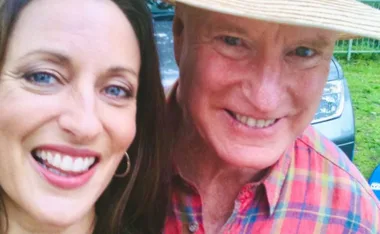 Smiling duo take a selfie outdoors, one wears a straw hat and plaid shirt, the other has hoop earrings.