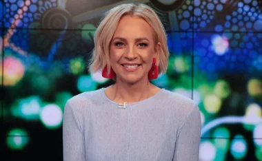 Blonde woman in red earrings and blue shirt smiles, with colorful abstract background.