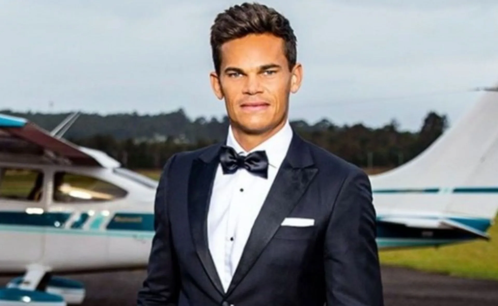 Man in a tuxedo standing in front of a small airplane on a runway.