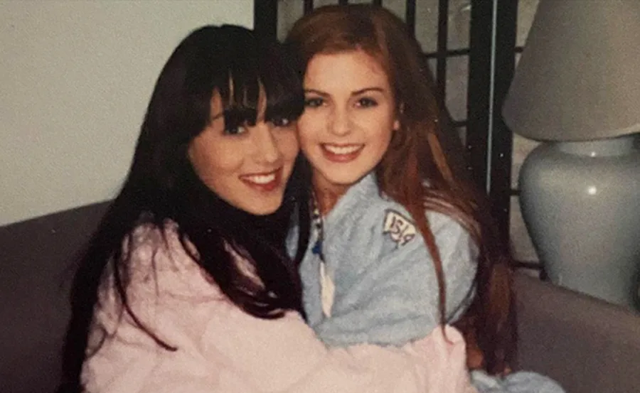 Two women embracing and smiling while sitting on a couch, both wearing casual wear.
