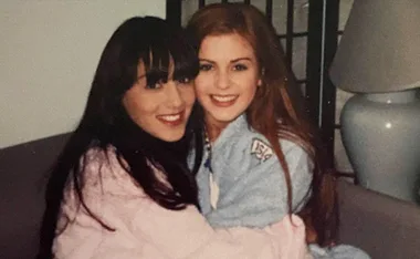 Two women embracing and smiling while sitting on a couch, both wearing casual wear.