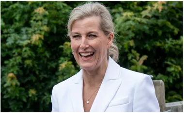 A woman in a white blazer smiles outdoors, with green foliage in the background.