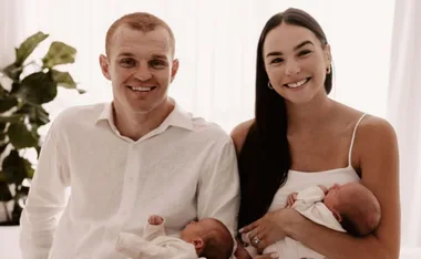 A smiling couple in white attire holds newborn twins, seated in a bright room with a plant in the background.