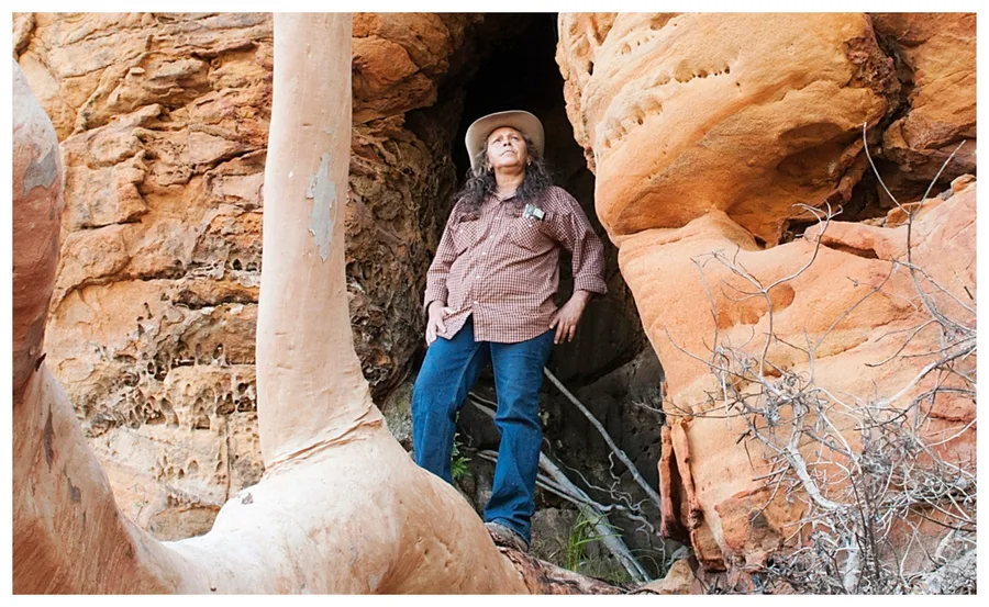 A woman in a cowboy hat stands confidently among large orange rock formations, looking up at the sky.