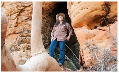 A woman in a cowboy hat stands confidently among large orange rock formations, looking up at the sky.