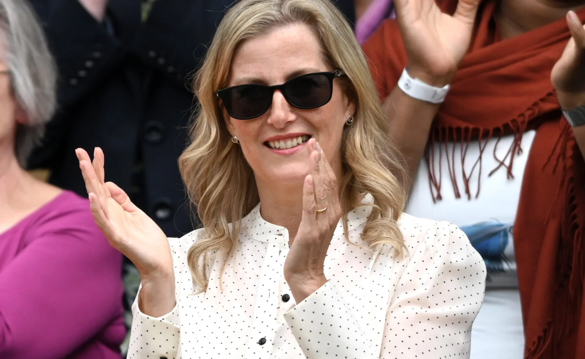 A woman with sunglasses and a polka dot blouse claps in a crowd at an outdoor event.