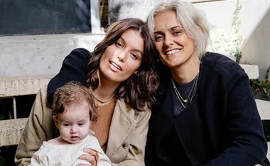 A smiling couple sits on steps with a baby, wearing casual attire, surrounded by greenery.