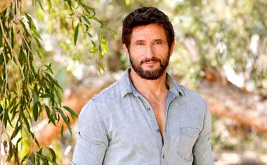 A man with a beard and short hair smiles outdoors, wearing a light denim shirt, with trees in the background.