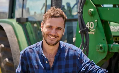 Man smiling in front of a green tractor wearing a blue checkered shirt outdoors.