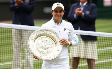 Tennis player holding Wimbledon trophy on court, wearing white outfit, with clapping crowd in background.