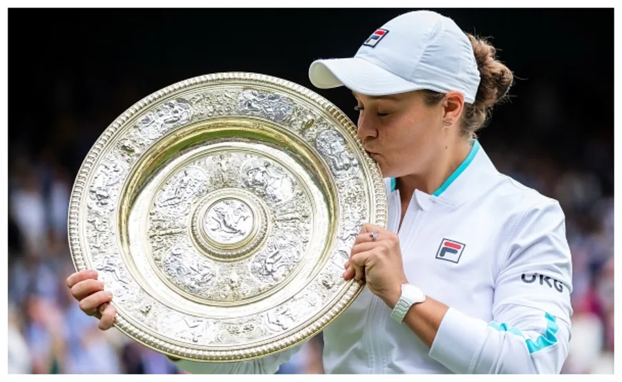Tennis player kissing the Wimbledon trophy on court after victory, wearing a white jacket and cap.
