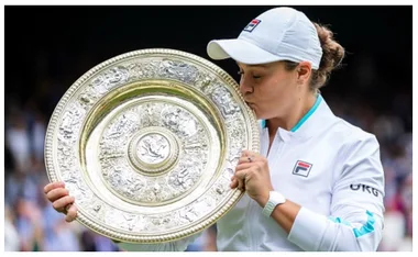 Tennis player kissing the Wimbledon trophy on court after victory, wearing a white jacket and cap.