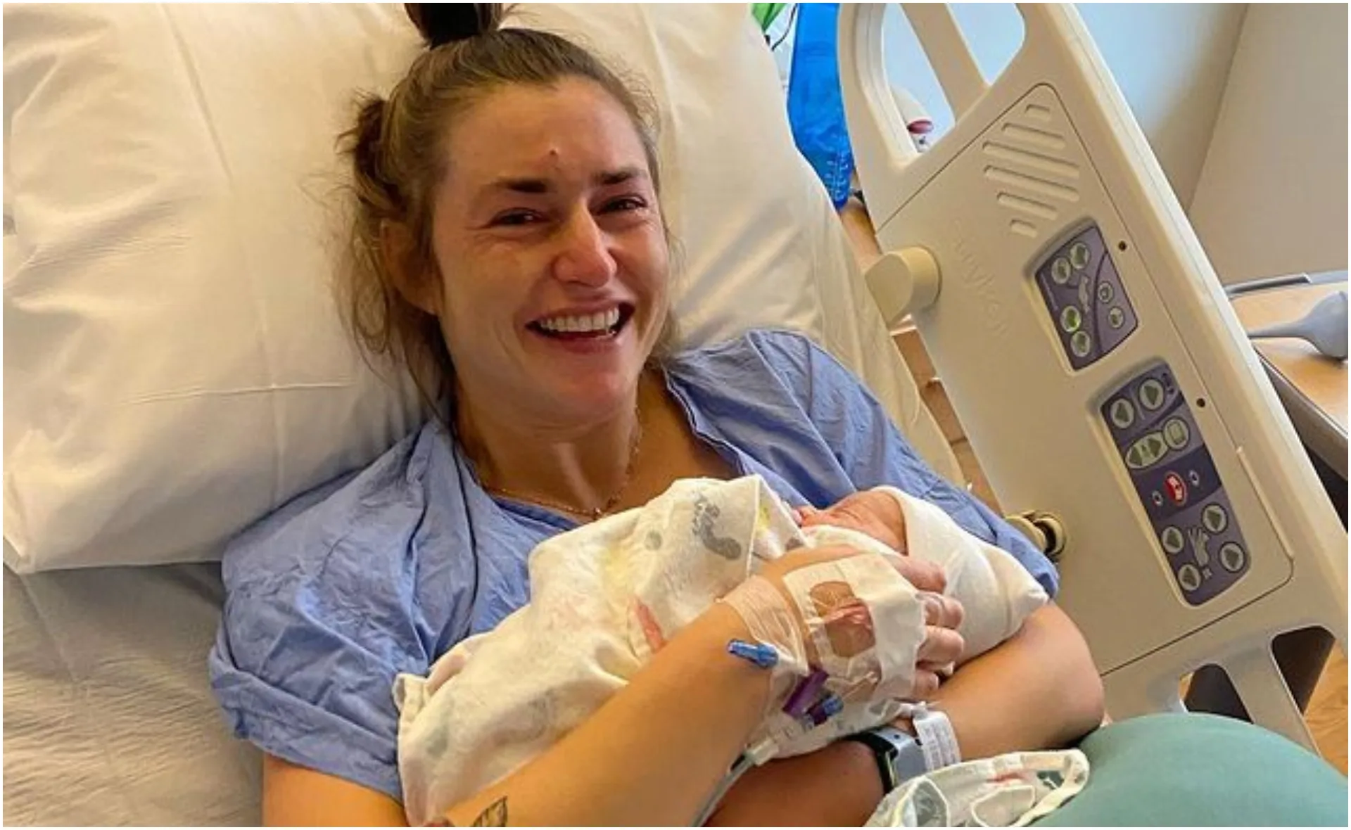 Woman in hospital bed smiling while holding newborn baby wrapped in a blanket.